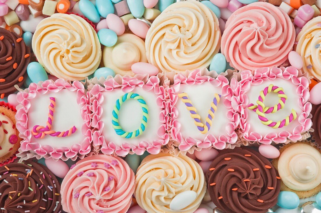 still life of four pink square iced cakes with letters on the top which spell the word love, surrounded by coloured cup cakes and sweets