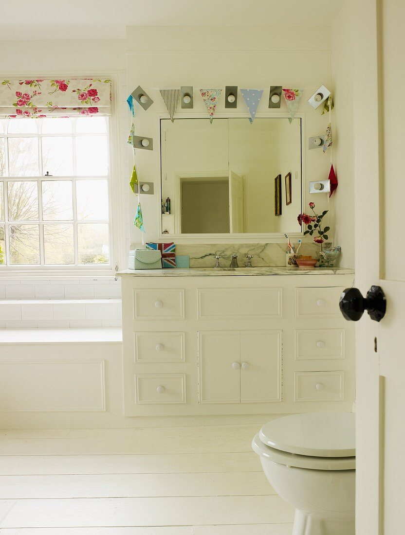 Bright bathroom with white furnishings, wall mirror surrounded by bunting and white wooden floor