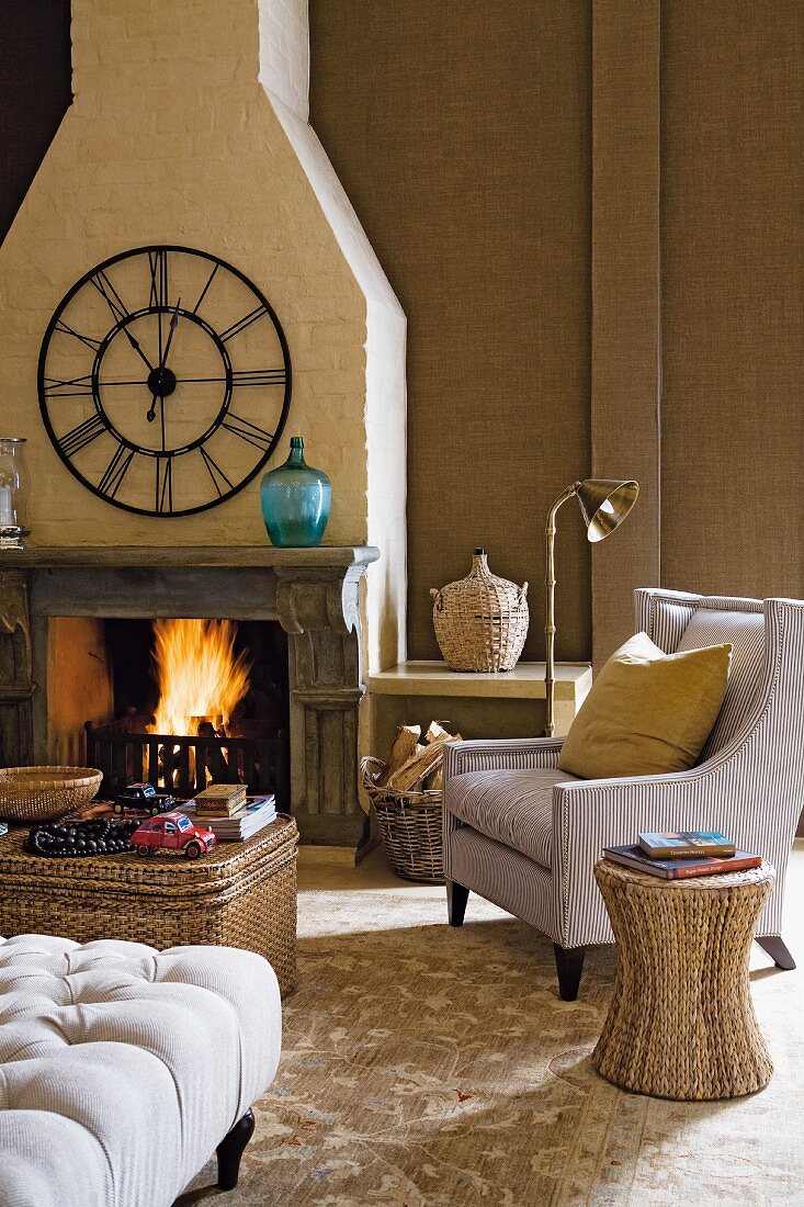 Rattan stool next to armchair and coffee table in front of fire in open fireplace below traditional, wrought iron wall clock