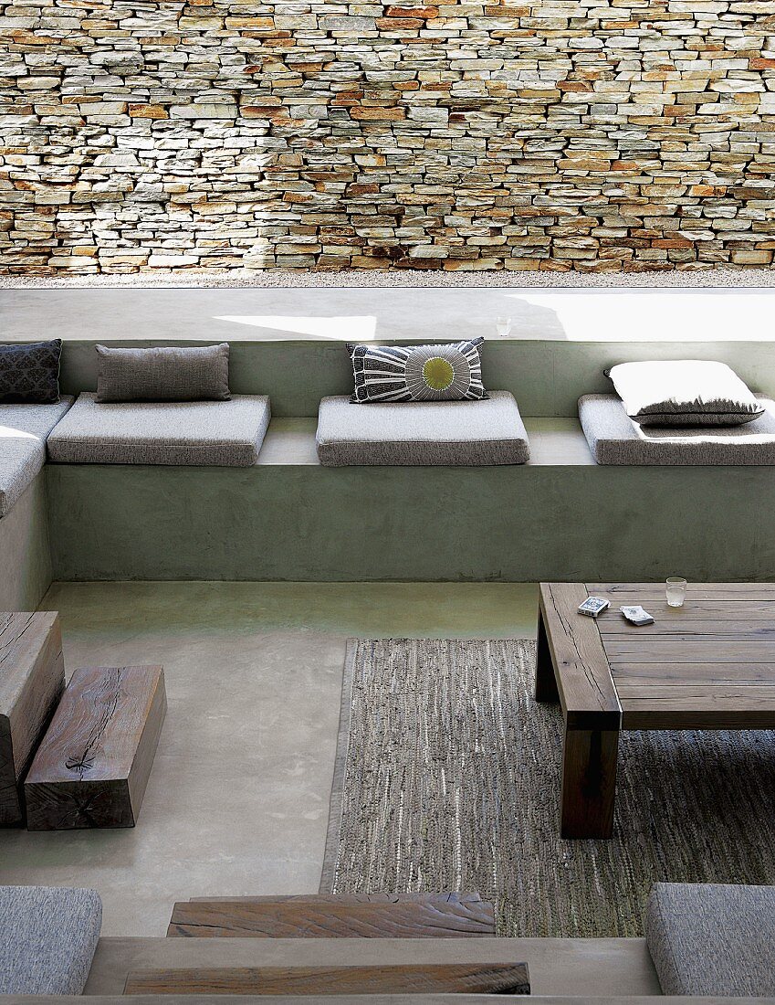 Sunken, concrete seating area with seat cushions and scatter cushions against stone wall