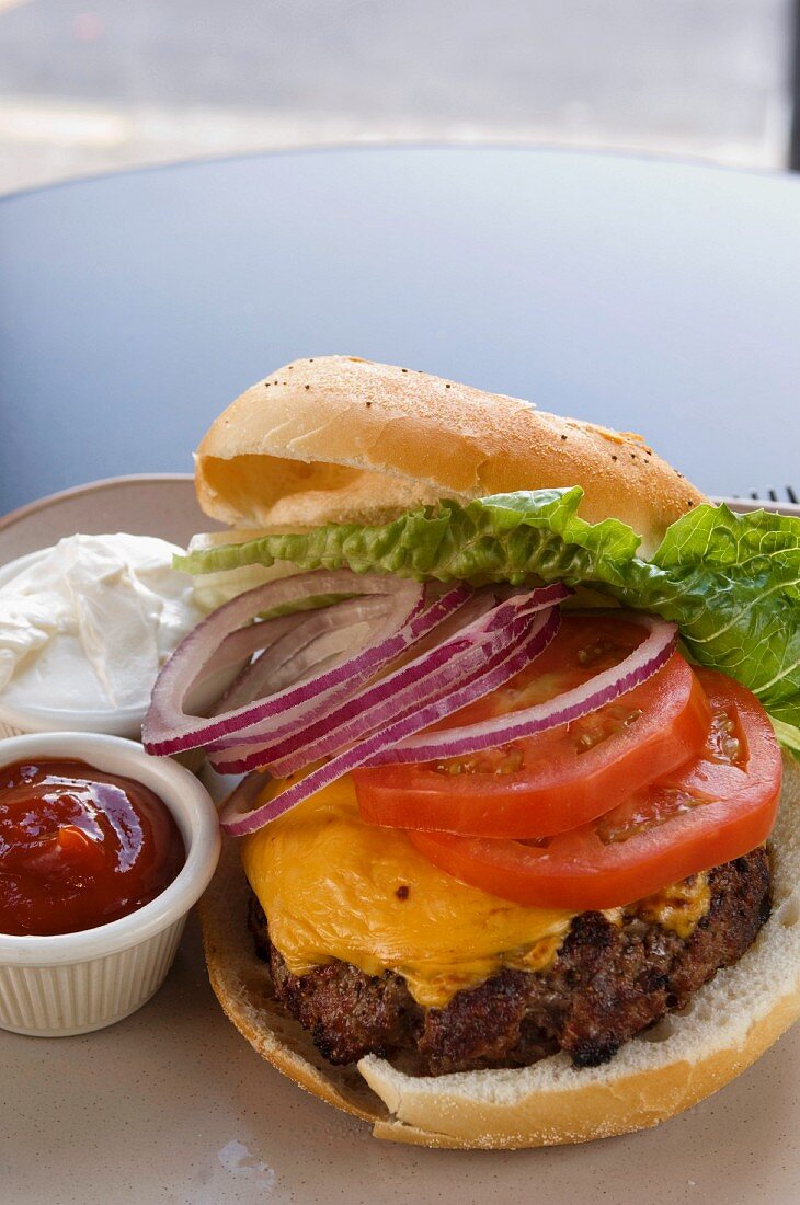 Cheeseburger mit Zwiebeln, Tomaten und Salatblatt, daneben Ketchup und Mayo