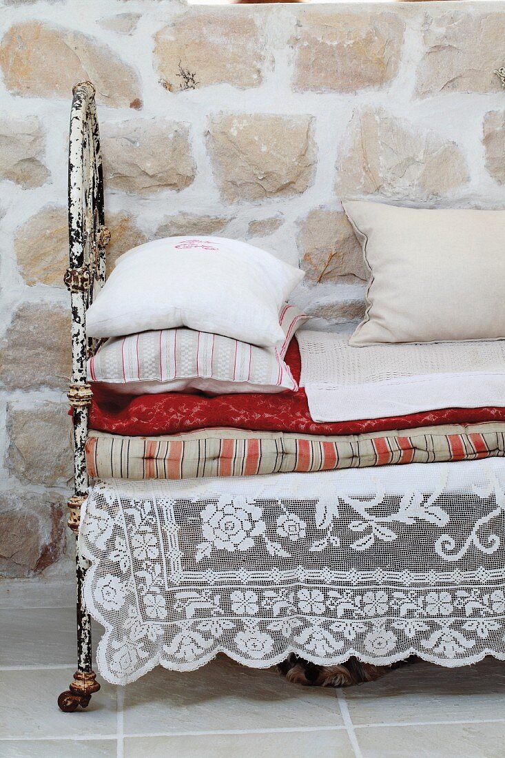 Lace cloth under stacked cushions and blankets on daybed with rusty metal frame against stone wall