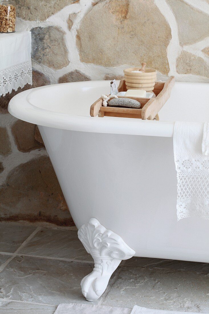 Toiletries on wooden rack over vintage, clawfoot bathtub in rustic bathroom with stone wall