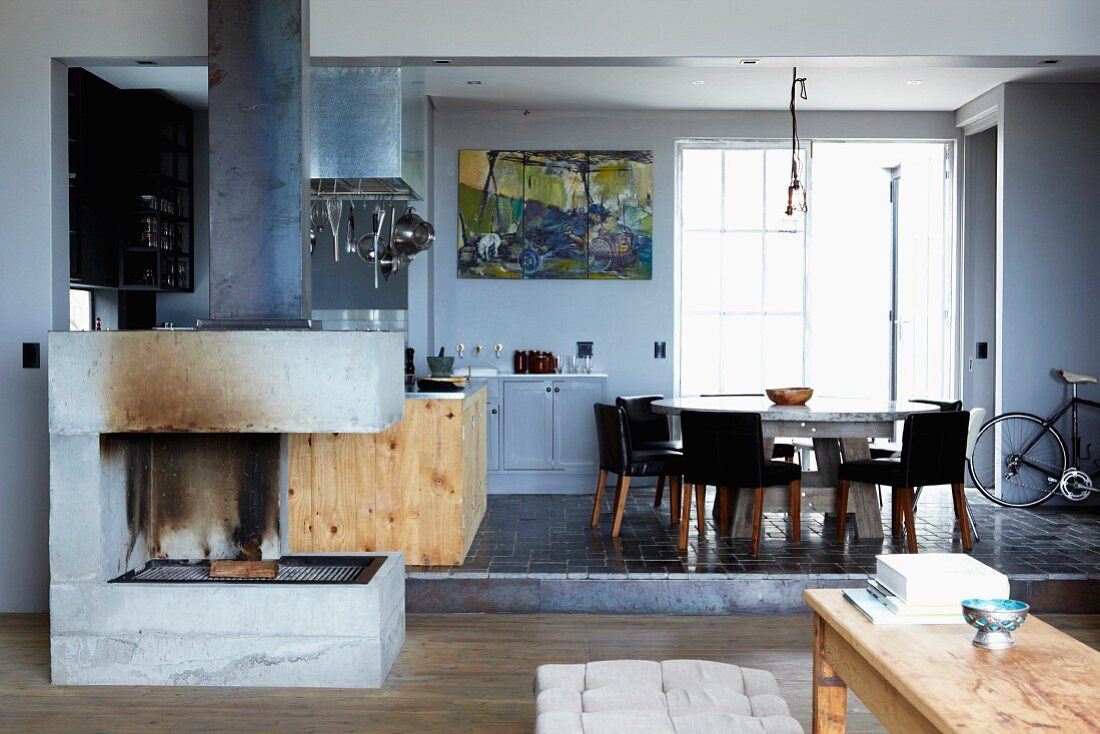 Concrete open fireplace in open-plan interior with dining table and chairs next to industrial window on black-tiled platform