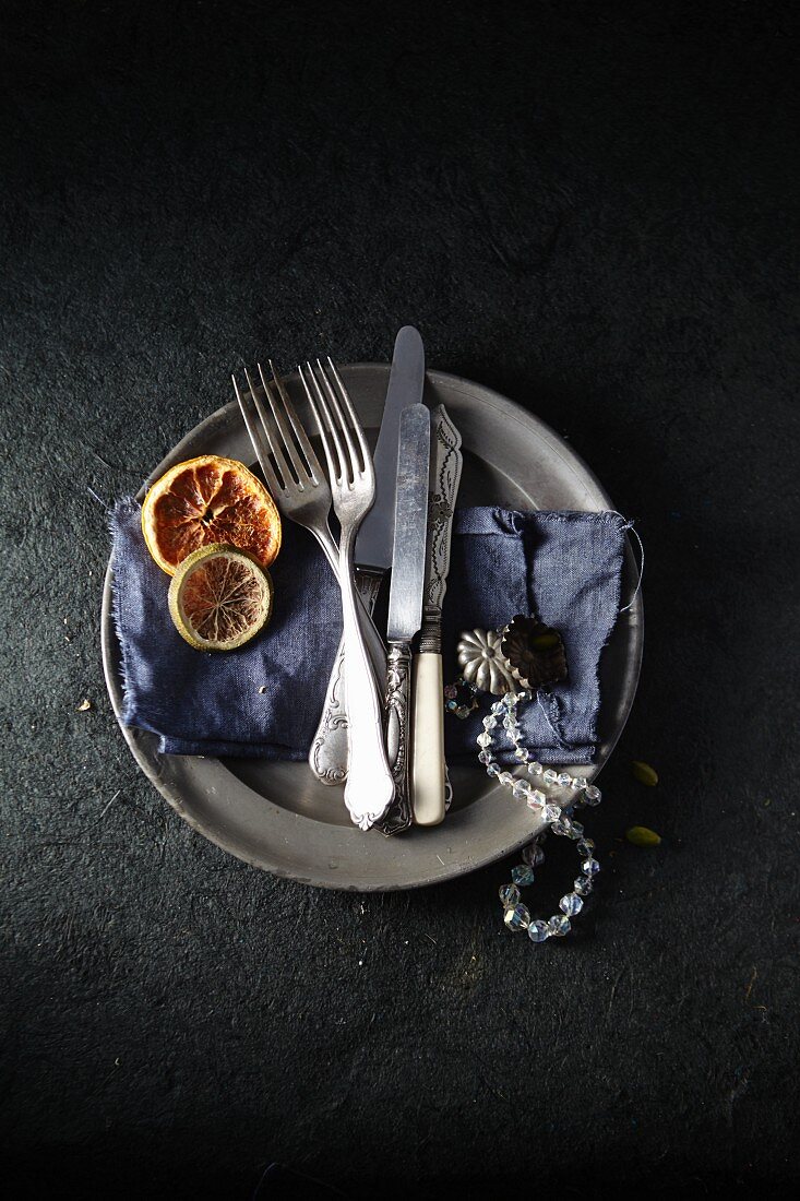 A tin plate with a blue cloth, antique cutlery, a necklace and dried citrus fruits