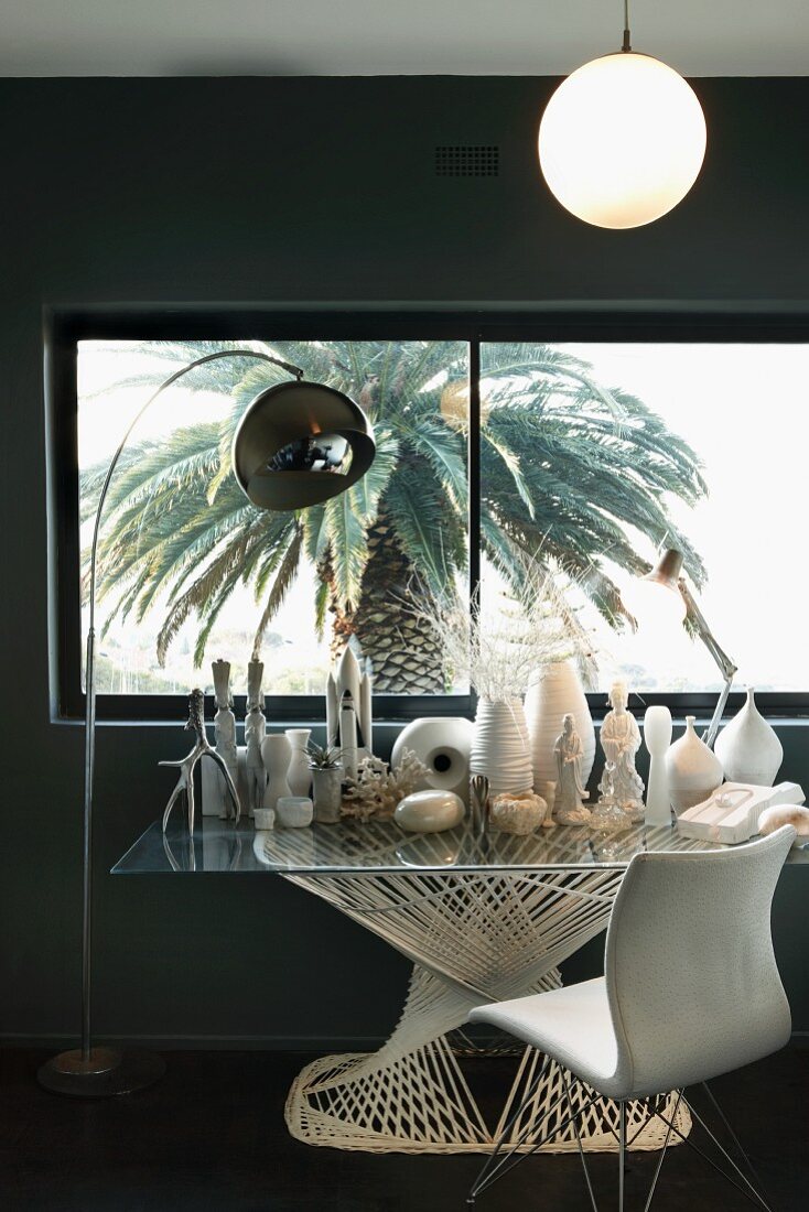 Collection of white vases and sculptures on modern glass table next to arc lamp and vintage chair in front of window with view of palm tree