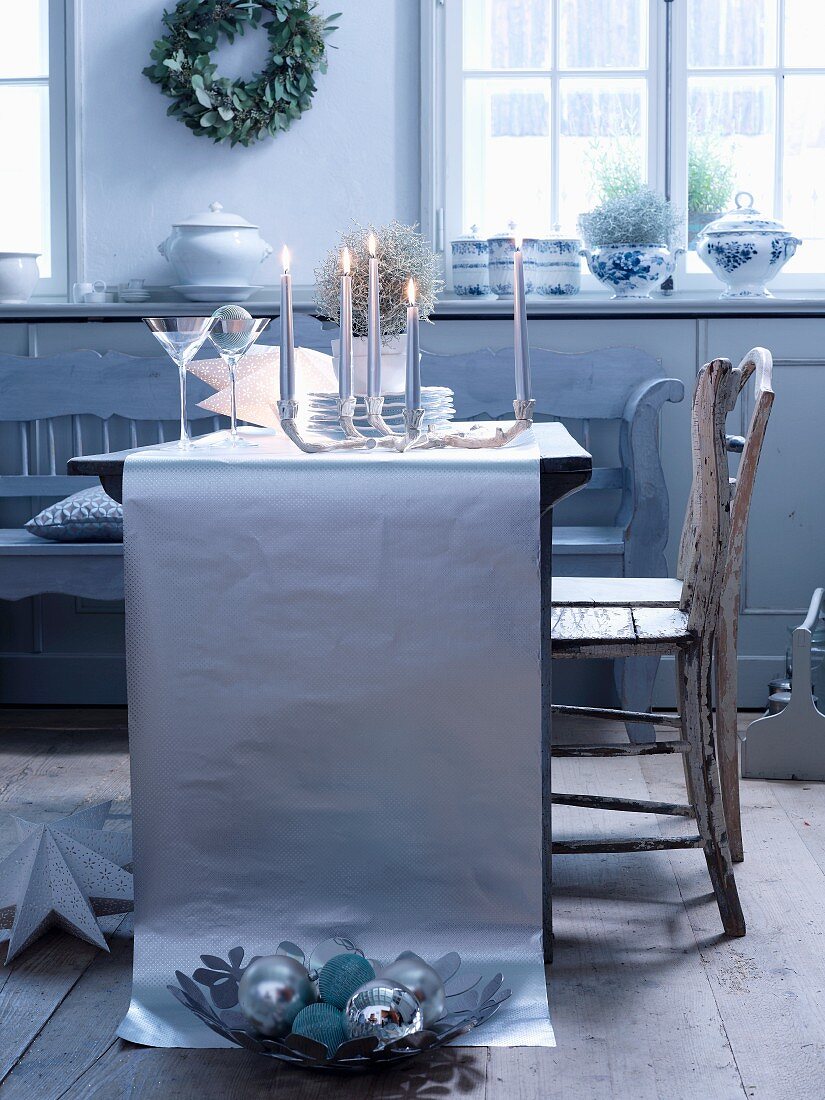 A table laid in silver - lit candles with glasses and plates on a table runner made from silver paper, with vintage wooden chairs pulled up to the table