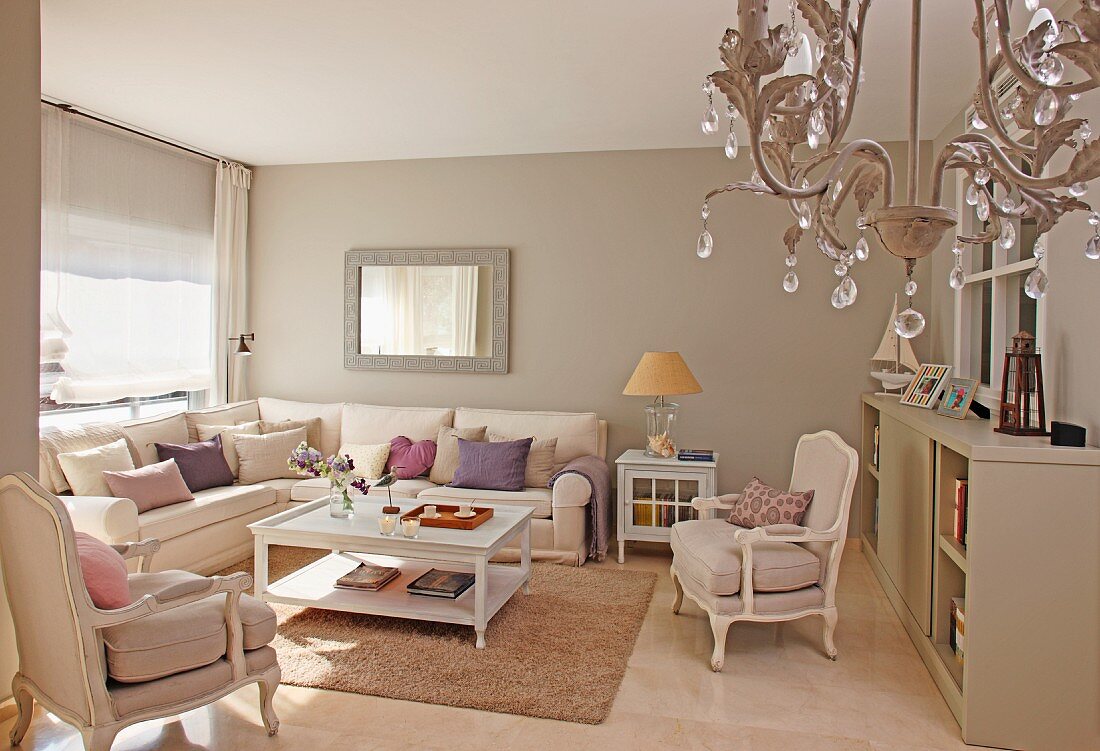 Living room in shades of beige with traditional corner sofa and antique-style armchairs; chandelier in foreground