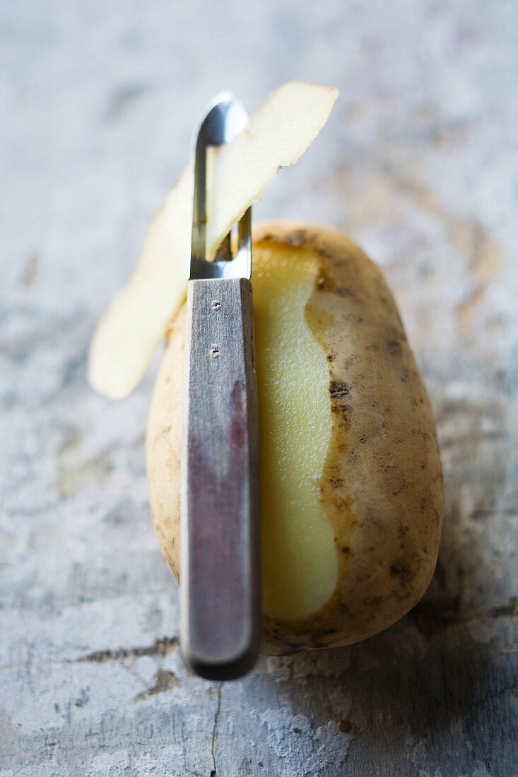 A potato, partly peeled, with a potato peeler