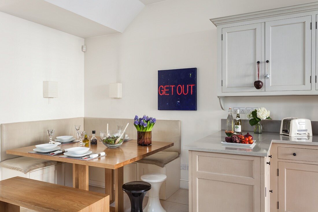 Blue-red typeface behind a square dining table with corner bench and simple kitchen with serene Shaker style color scheme