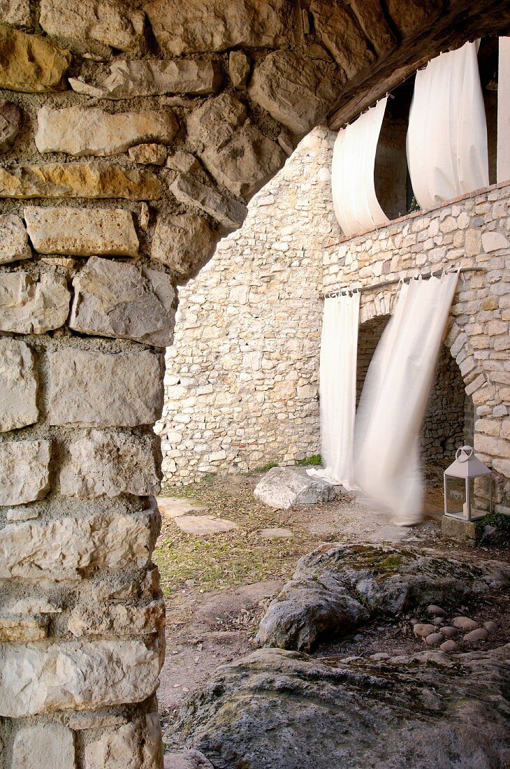 View through stone arch into courtyard with arched doorway and balcony with white curtains