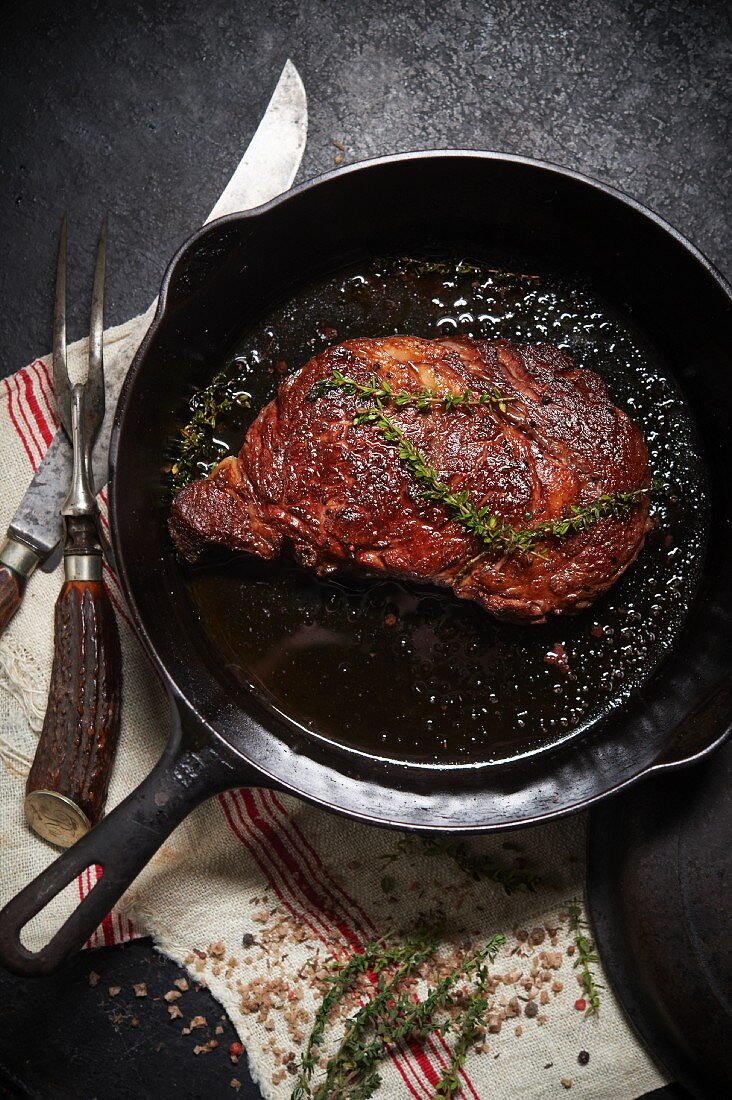 Ribeye Steak in einer Gusseisenpfanne mit Thymian, Fleischgabel und Messer