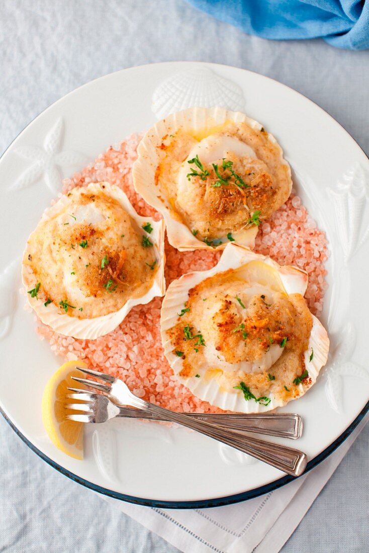 Baked Scallops with Cheese and Wine Sauce on Pink Salt on a White Plate; Forks and Lemon