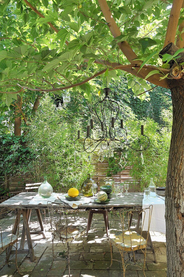 Rustikaler Tisch mit Vintage Metallstühlen unter Baum mit aufgehängtem Kerzenleuchter in mediterranem Garten