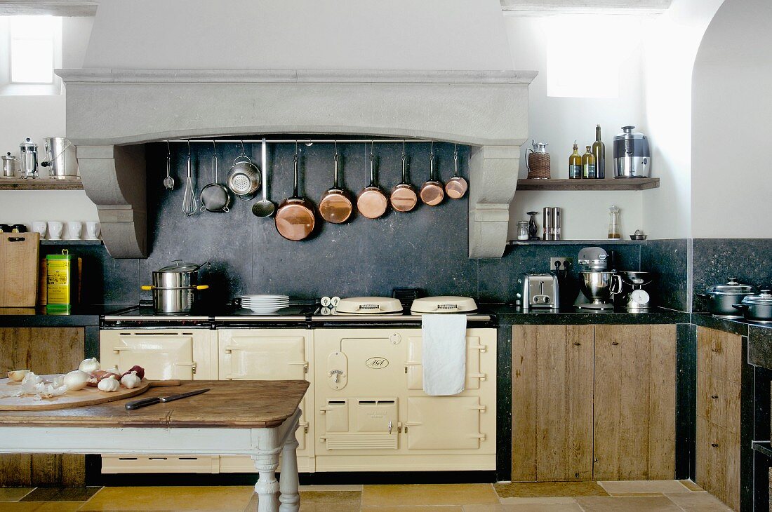 Antique kitchen table with chopping board on table in front of vintage range and collection of copper pans hung in masonry extractor