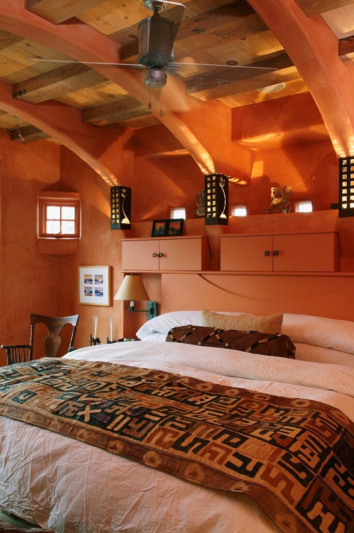 Attic bedroom with exposed wooden roof structure - double bed against salmon pink wall with small cupboards on headboard in same colour