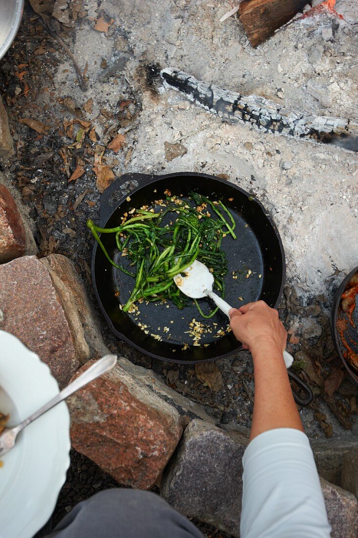 Removing Sauteed Rapini Out of an Iron Skillet Over a Fire Pit