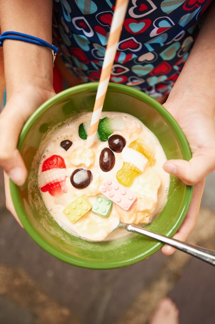 Person Holding a Bowl of Melting Ice Cream with Candies, a Straw and a Spoon