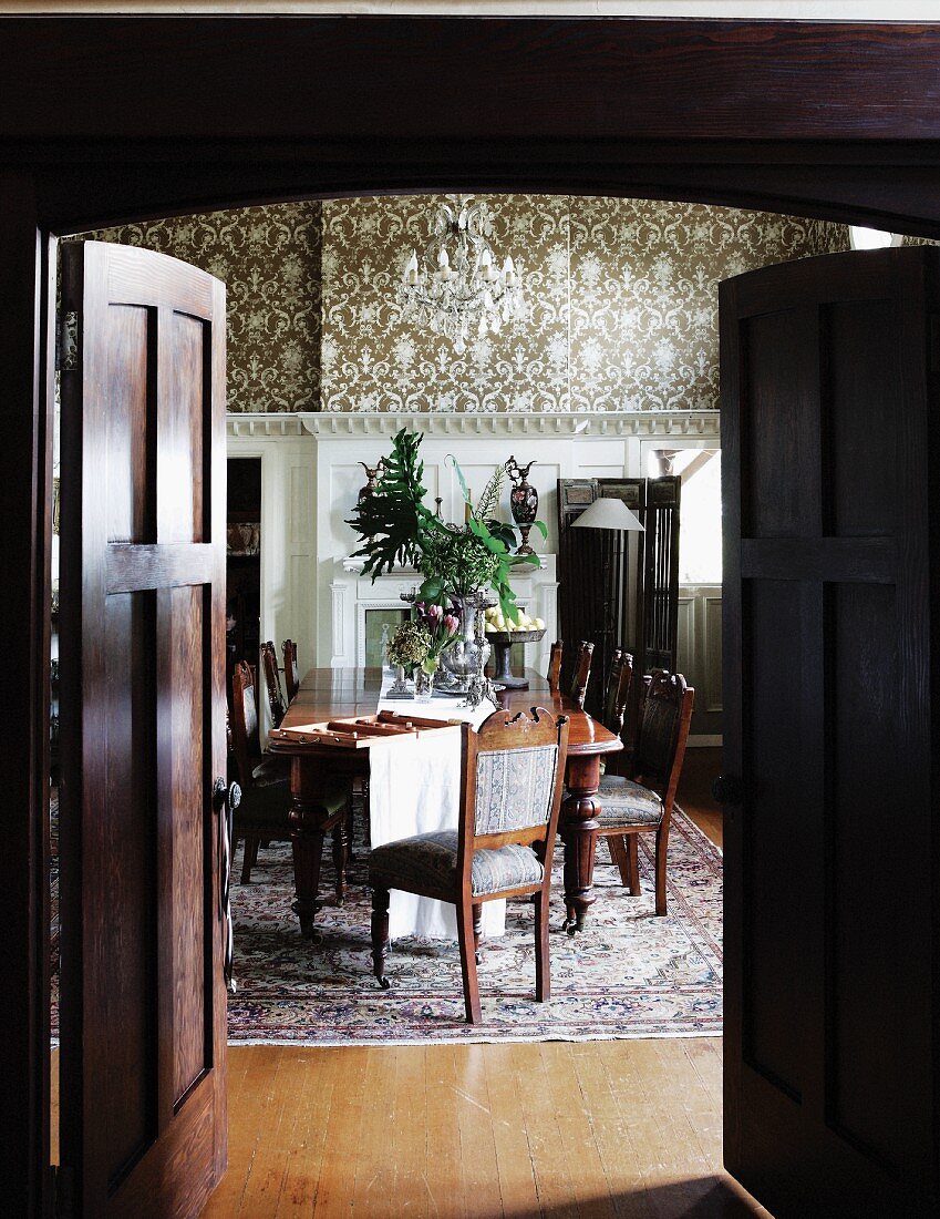 View through a dark wooden double door into a traditional dining room with a long dining table and colonial style, antique chairs