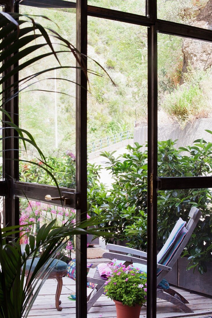 View of sun lounger on wooden terrace surrounded by foliage plants through open French windows
