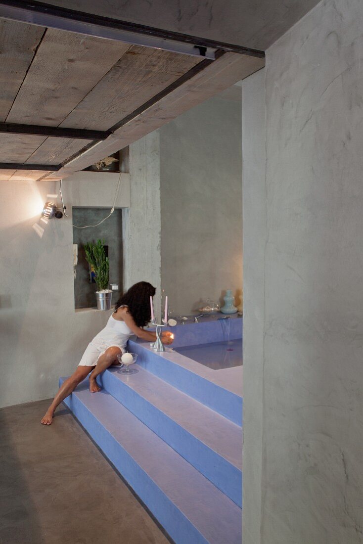 Exposed concrete walls and polished concrete floor contrasting with lavender stepped platform leading to sunken bathtub; woman lighting candles on edge of bath