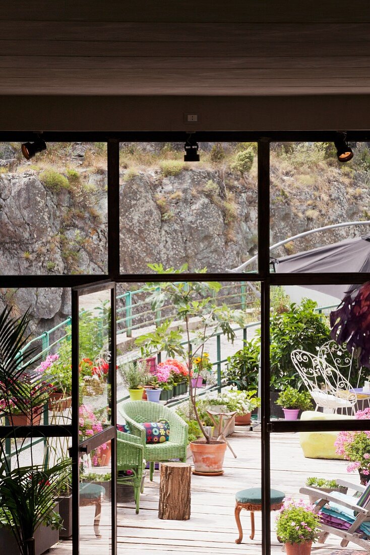 View through open door in floor-to-ceiling, industrial glazing onto platform-style terrace with chairs and potted plants