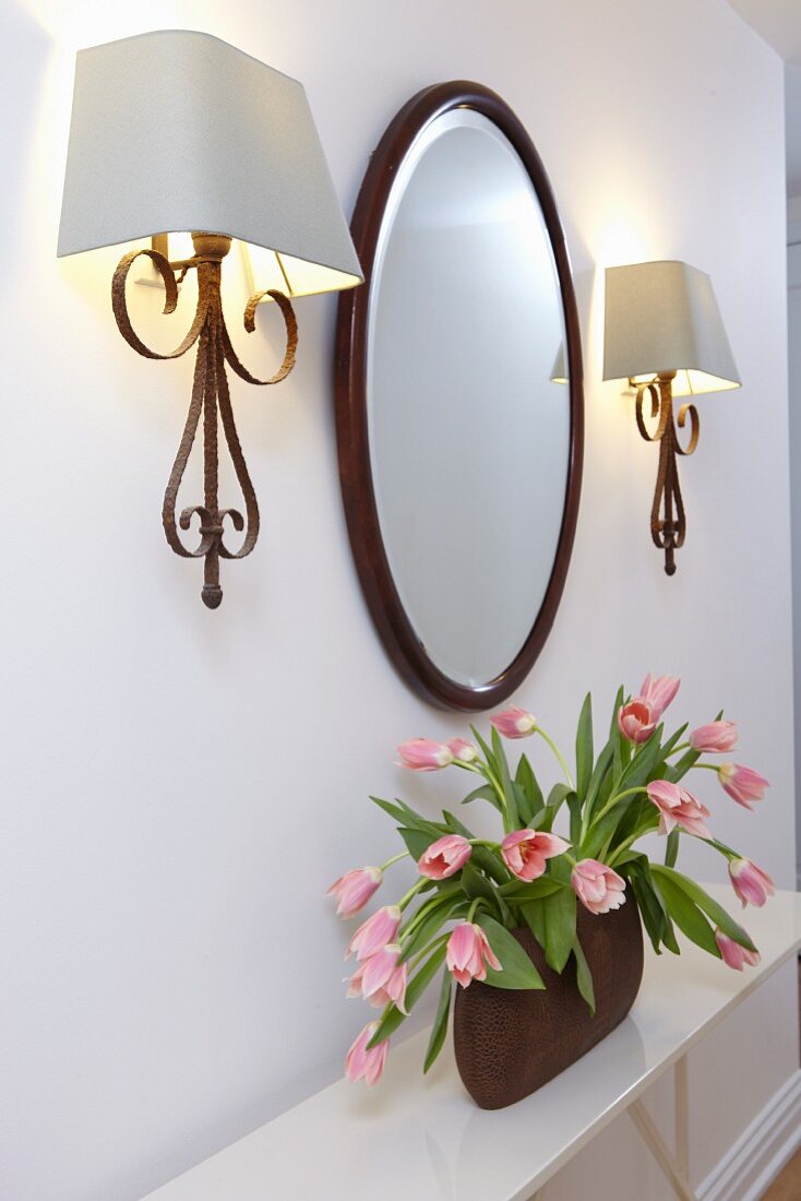 Mirror flanked by vintage sconce lamps with white lampshades above bouquet of tulips on sideboard