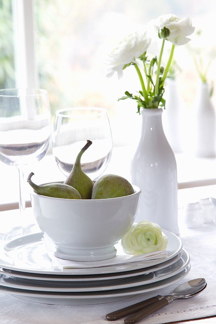 A stack of crockery with figs and a vase of white ranunculus flowers