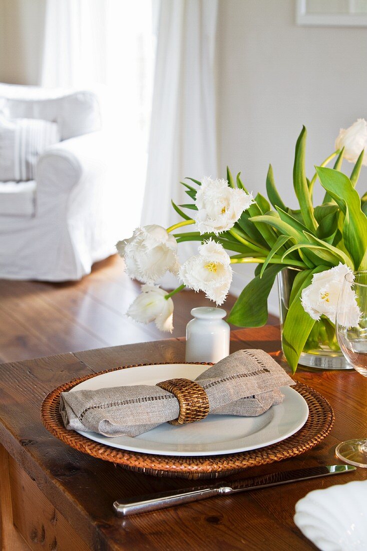 A bunch of white tulips in a glass vase; a place setting with a basket plate to the front of the image