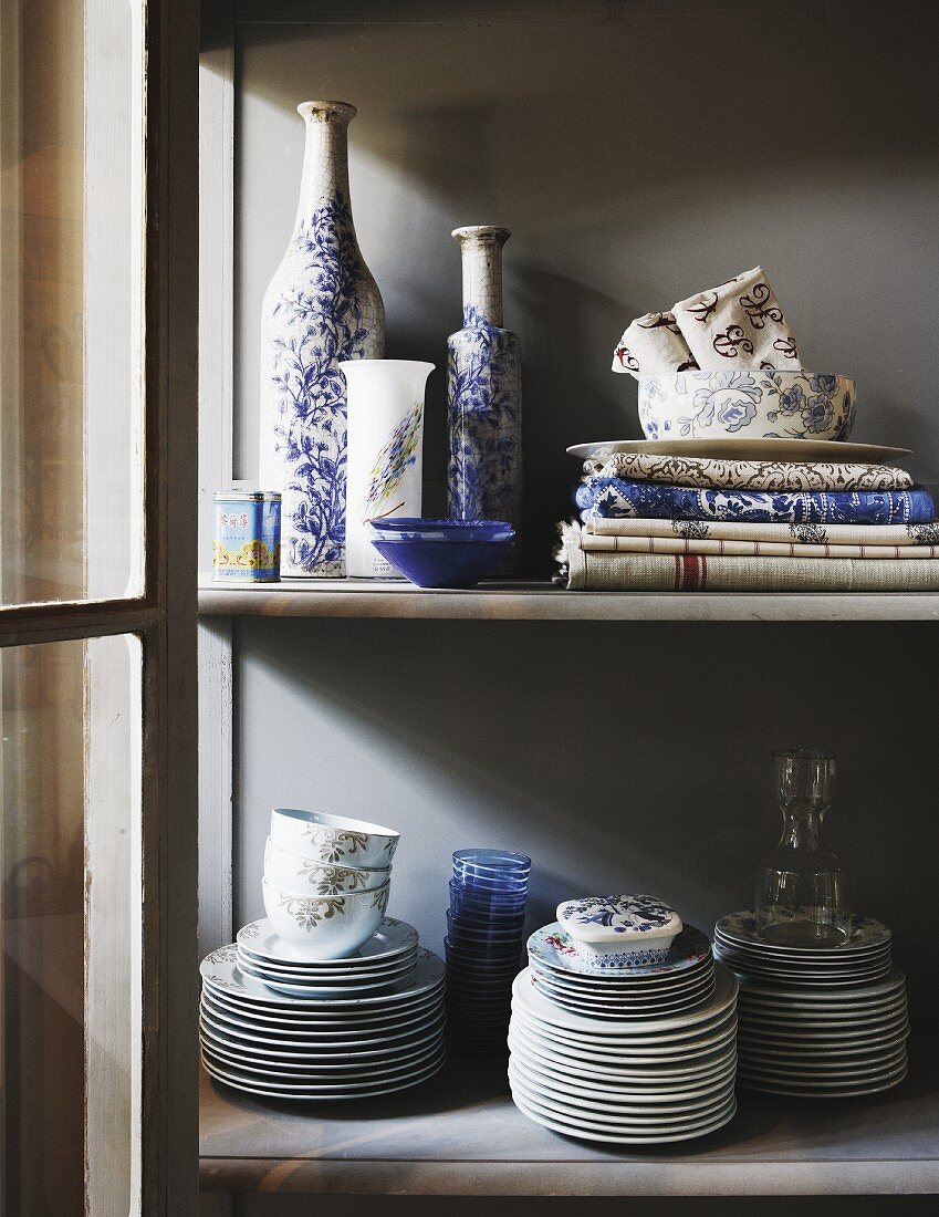 Display case with open door and view of white and blue painted bottles next to tablecloths and several stacks of plates