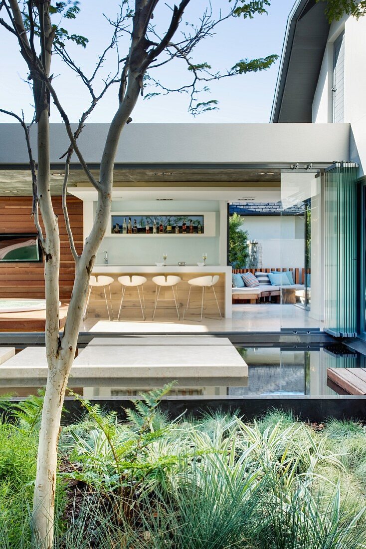 View across a planting bed and water feature through open folding doors into a one story building with a kitchen bar