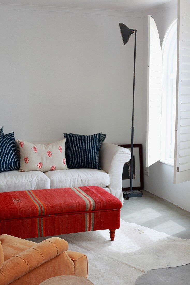 Coffee table covered in ethnic fabric in front of a light sofa and retro floor lamp in a minimalist room with interior shutters at the arched window