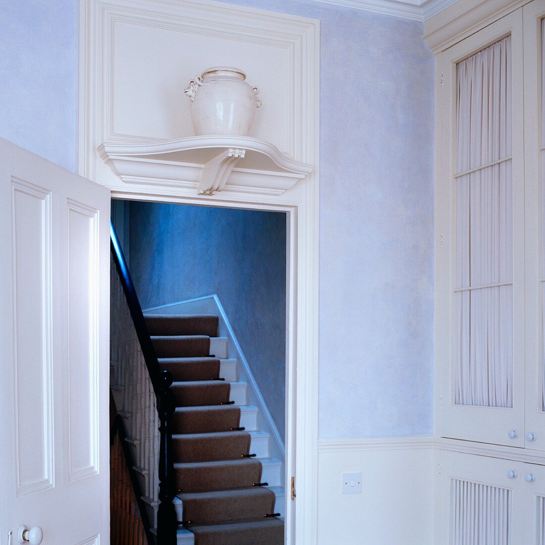 Antique urn on white, carved wooden bracket above open interior door showing staircase with runner