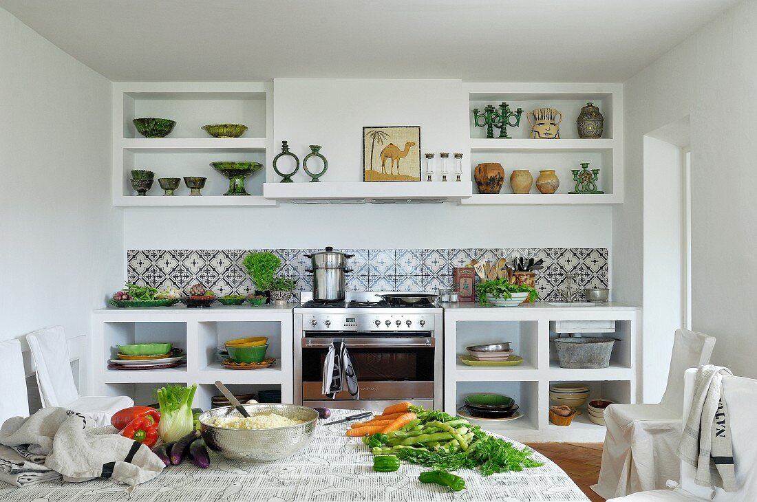 Weiße Küchenzeile mit Keramikfliesen; im Vordergrund ein großer Tisch mit frischem Gemüse und einer Edelstahlschüssel voller Couscous
