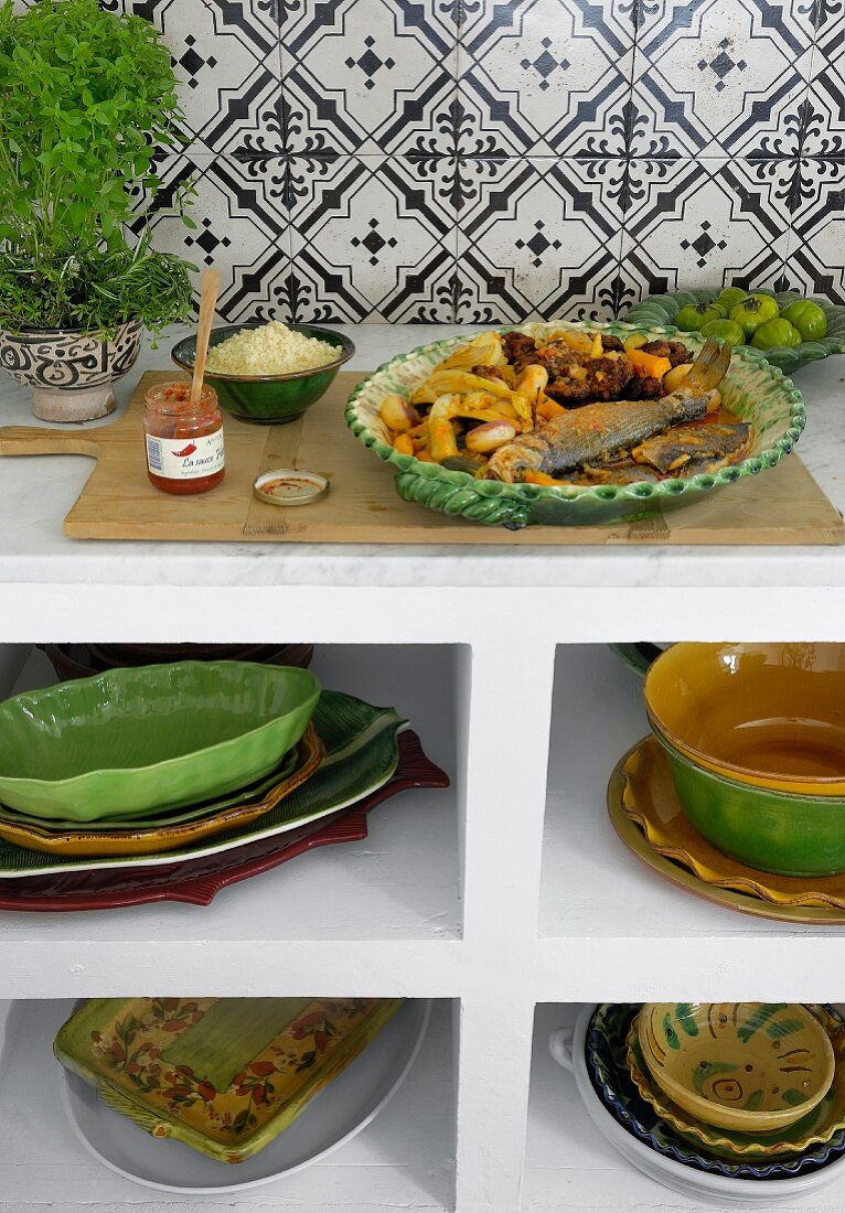 Küchenregal mit gestapeltem Keramikgeschirr; darauf eine Platte mit Fisch und Gemüse aus dem Ofen und ein Glas Harissasauce