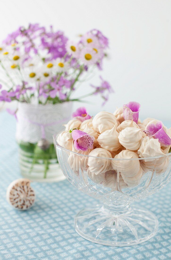 A Glass Footed Bowl Full of Meringue Cookies with Flower Petals; Vase of Flowers in Background