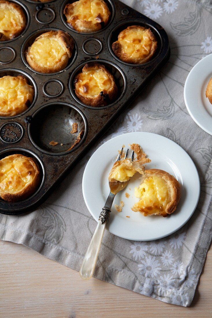 Portuguese Custard Tarts in a Baking Pan and on a Small White Plate with a Fork; From Above