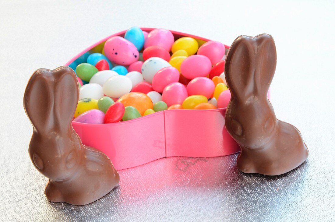A heart-shaped tin of colourful sugar eggs; to the front, two chocolate bunnies