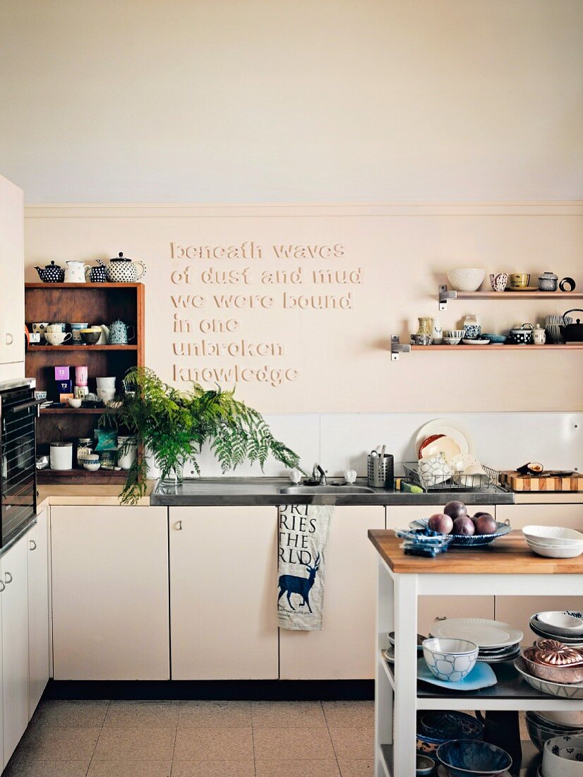 Wandboarde und Regale mit zahllosem Keramikgeschirr und Sinnspruch-Relief an Küchenwand