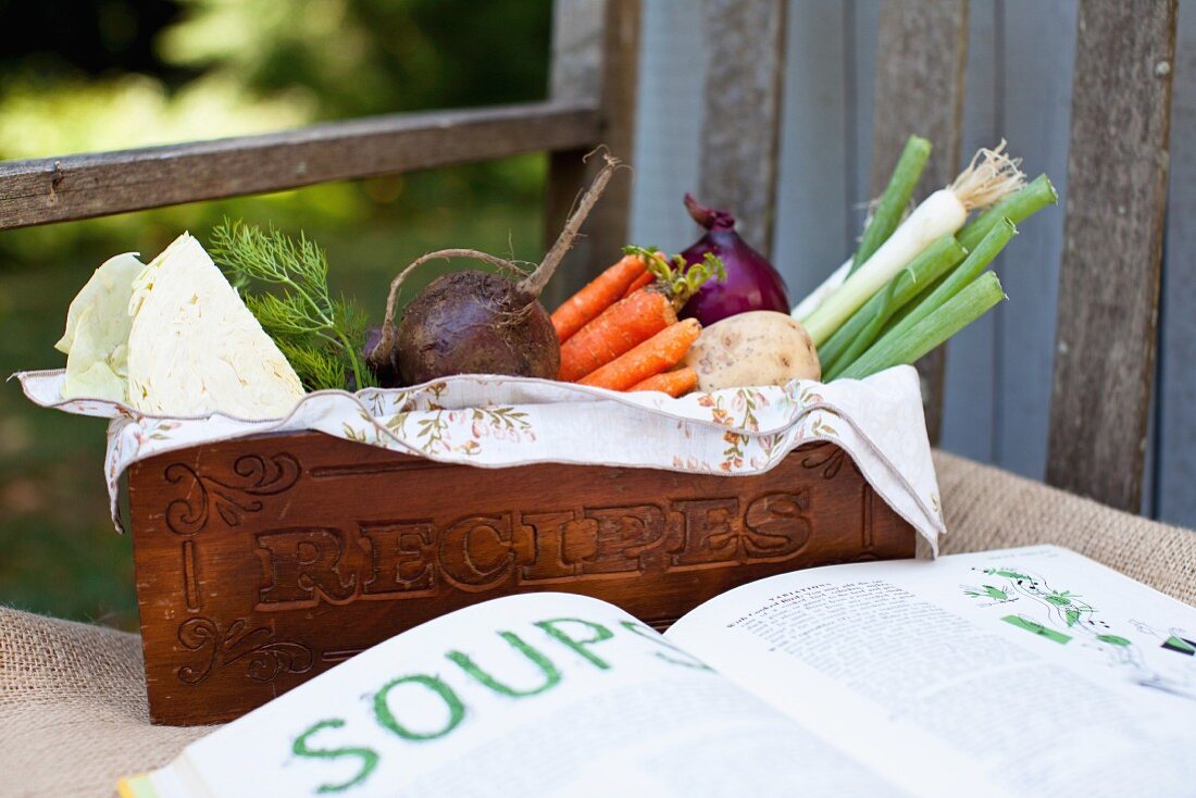 Frisches Gemüse aus dem Garten in einer Holzkiste und ein aufgeschlagenes Kochbuch
