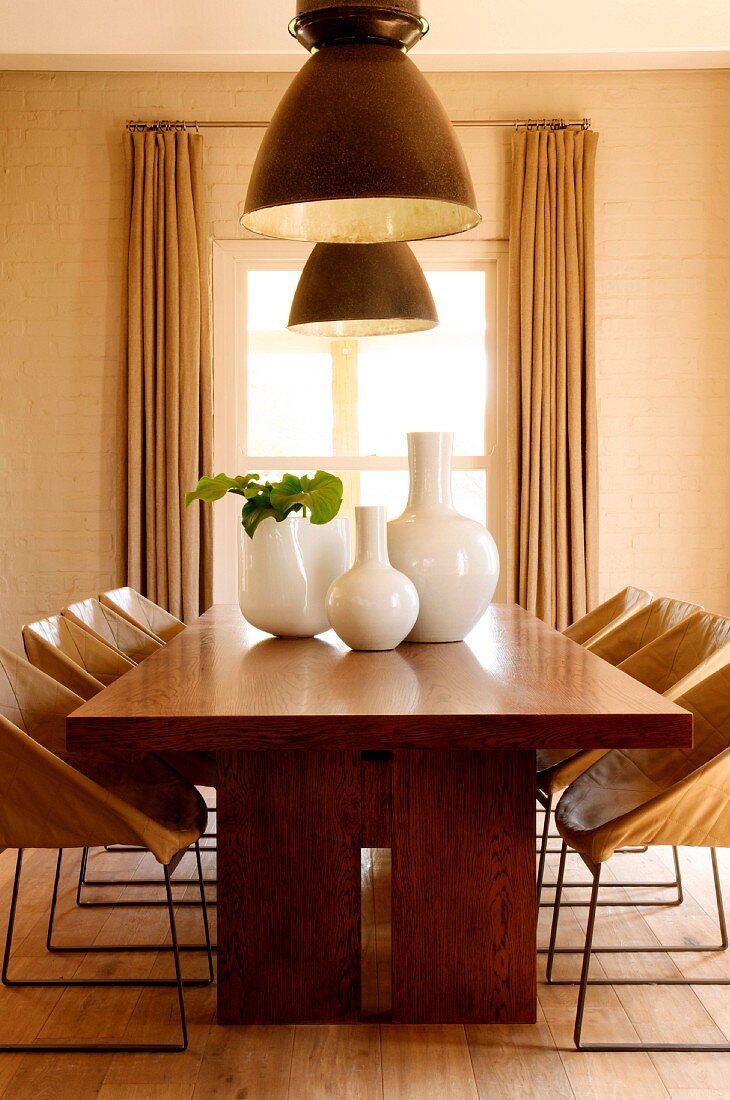 White ceramic vases on solid wood dining table below large ceiling lamps in front of dining room window