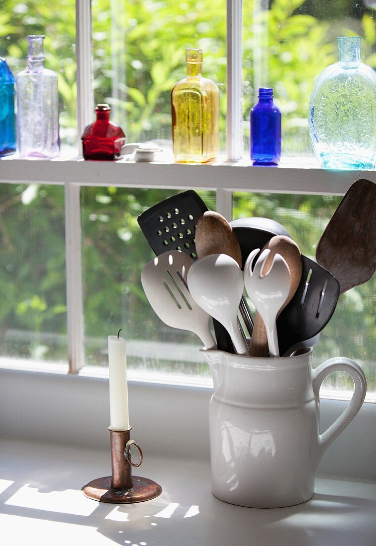 Assorted Kitchen Utensils in a Crock by a Window; Glass Bottles on the Windowsill