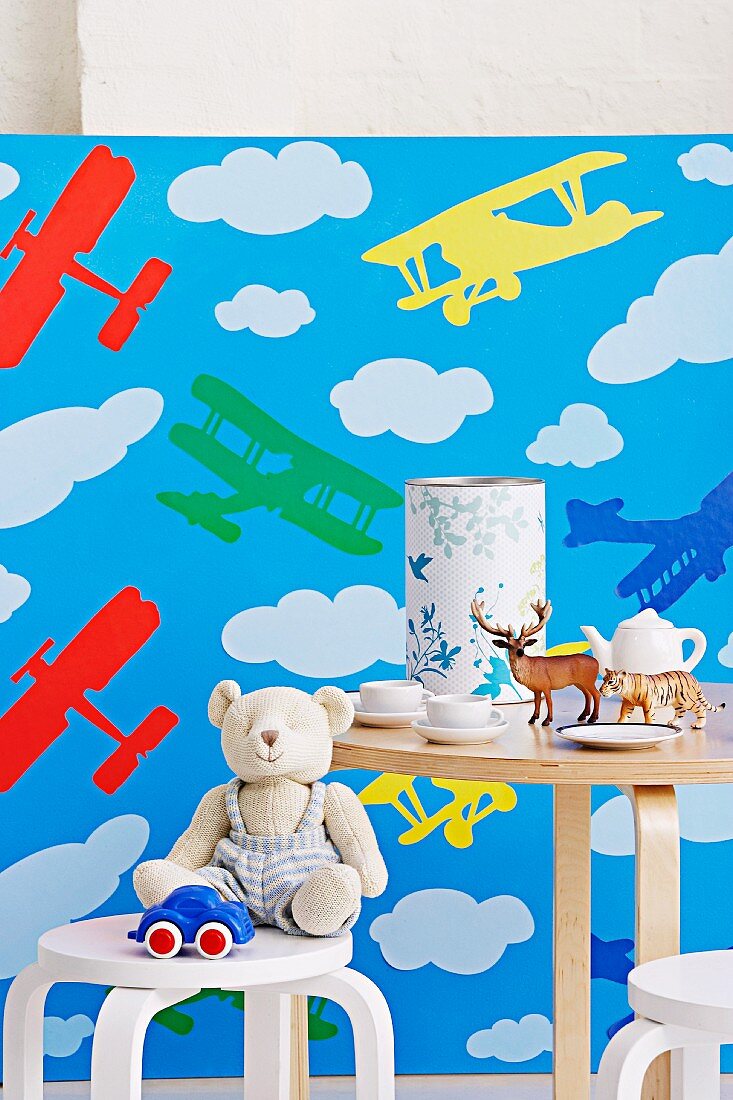 Kinderstuhl und Tisch mit Spielsachen, Kindergeschirr und Spardose vor Tapete mit Flugzeugmotiv