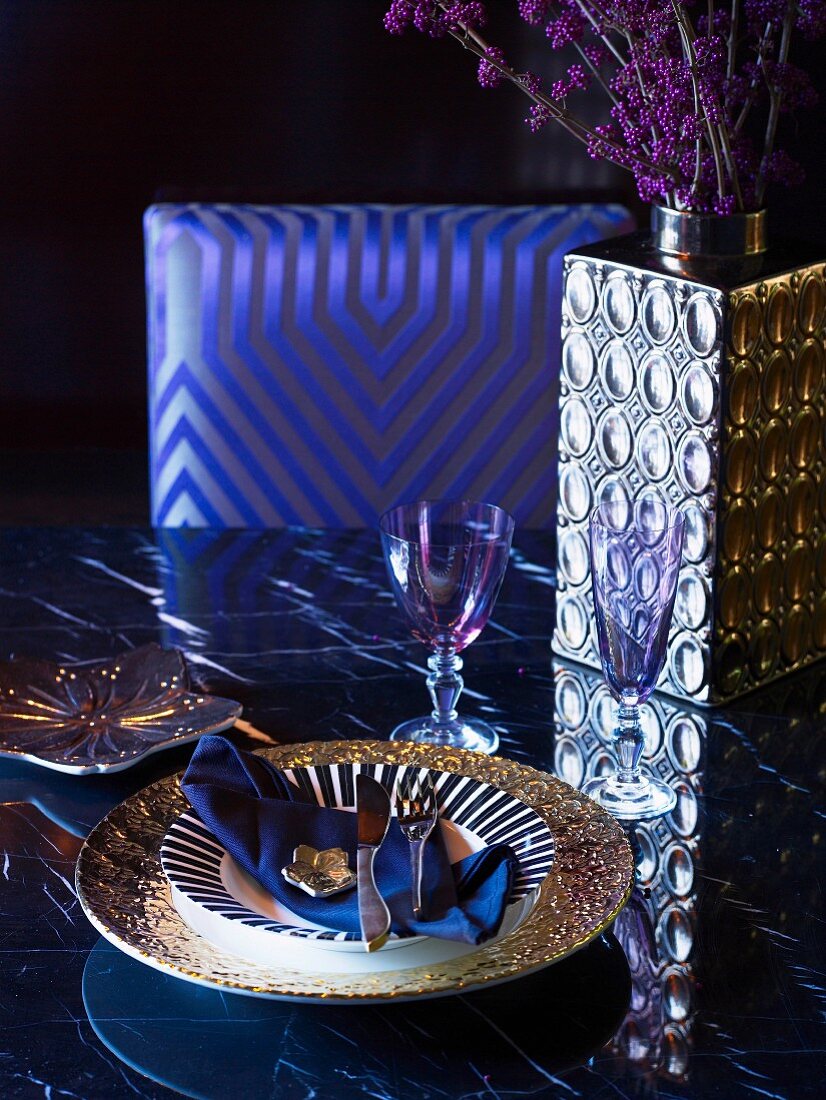 Place setting with gold-rimmed plate and shiny silver vase on dining table in front of chair with elegant upholstered backrest