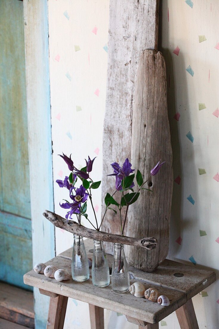 Clematisblüten in drei Glasfläschen mit Treibholzast arrangiert, dahinter Treibholzstücke auf Holzhocker in Vintageambiente
