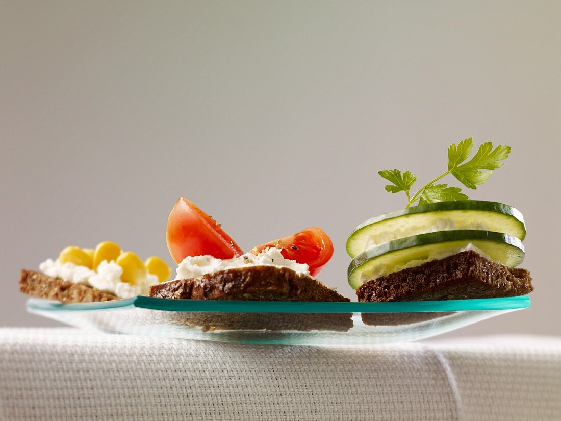 Three slices of bread topped with cottage cheese and with sweetcorn, tomato and cucumber respectively