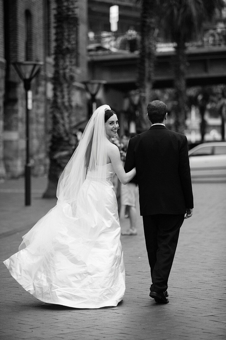 Bride wearing long white dress and groom in black suit walking down the street (black and white photo)