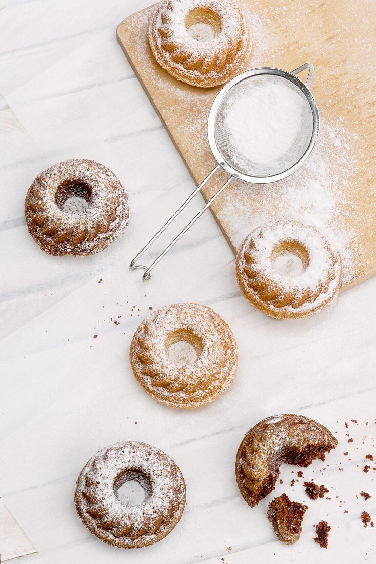 Mini Bundt cakes on grease-proof paper and on a wooden board with a sieve and icing sugar