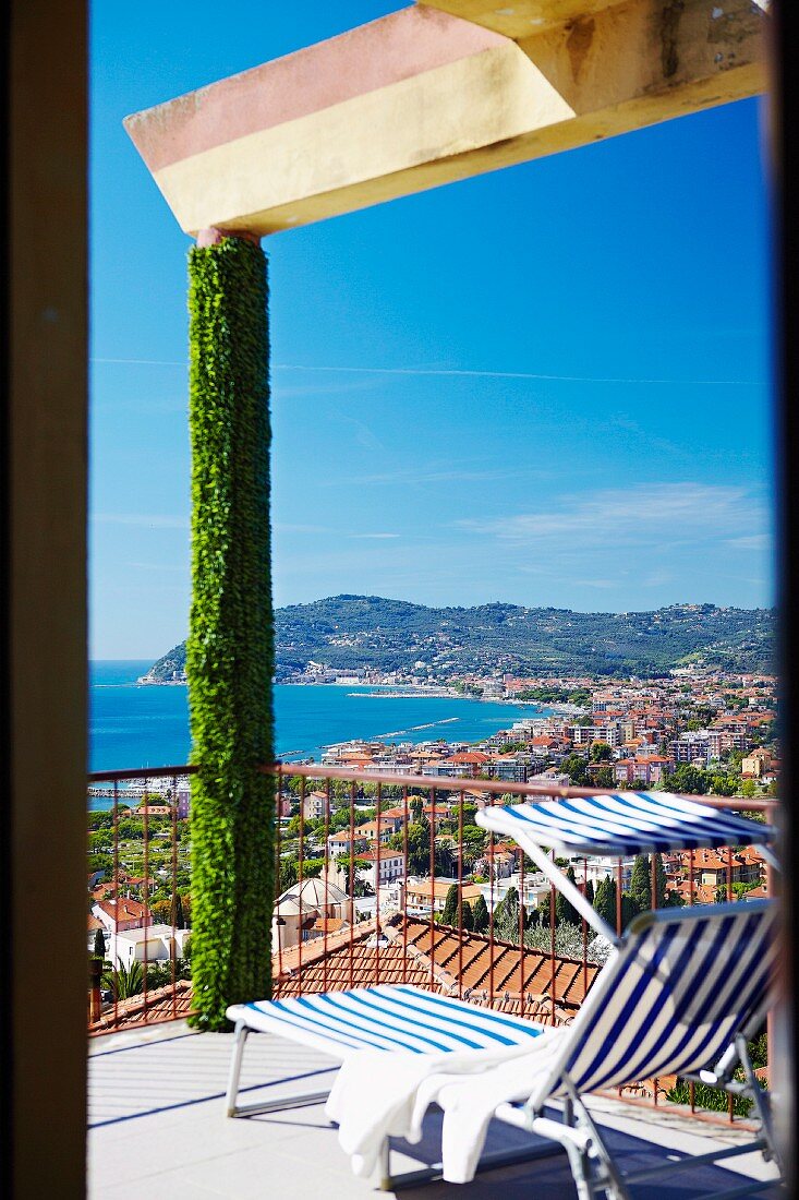 Blue and white striped sun lounger on terrace with climber-covered pillar; view over bay on Italian Riviera