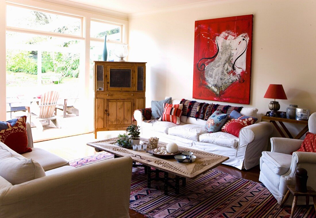Wooden furnishings, sofas with scatter cushions and striking red painting in interior with view out onto sunny terrace